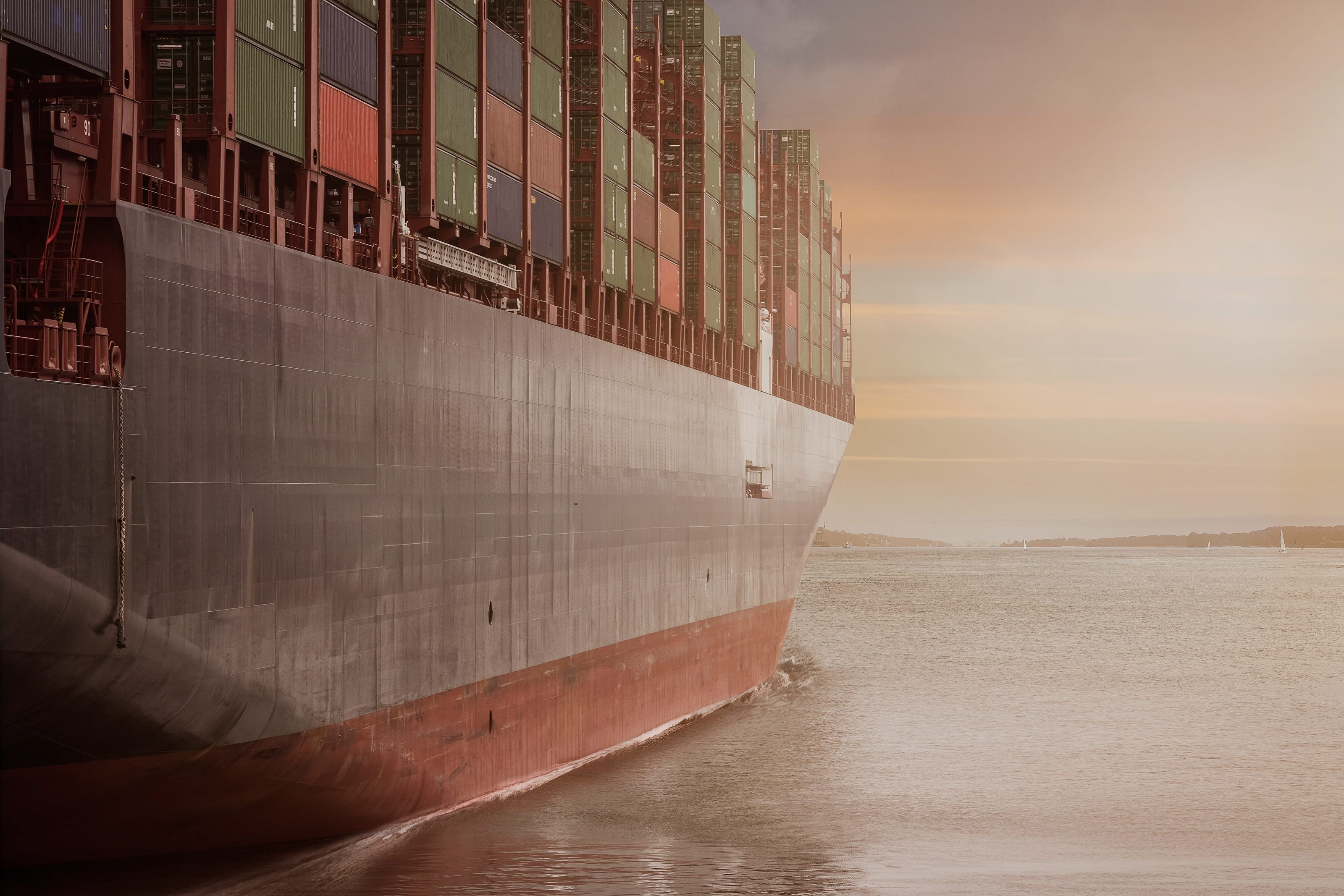 Hull of a fully loaded container ship moving through calm water at dusk