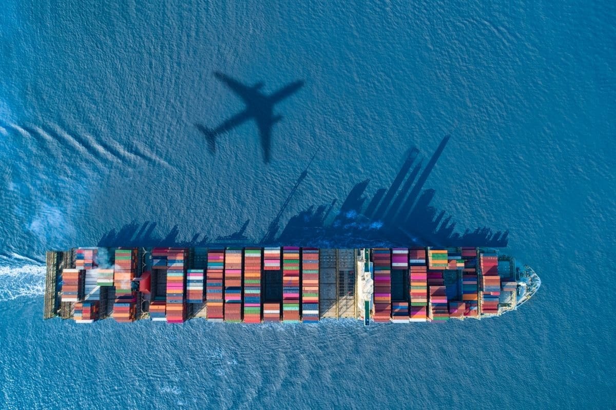 Top down view of a loaded container ship at sea with the shadow of an aircraft cast across the water
